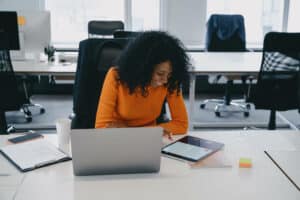 woman sitting down in front of laptop on her desk while looking at her tablet