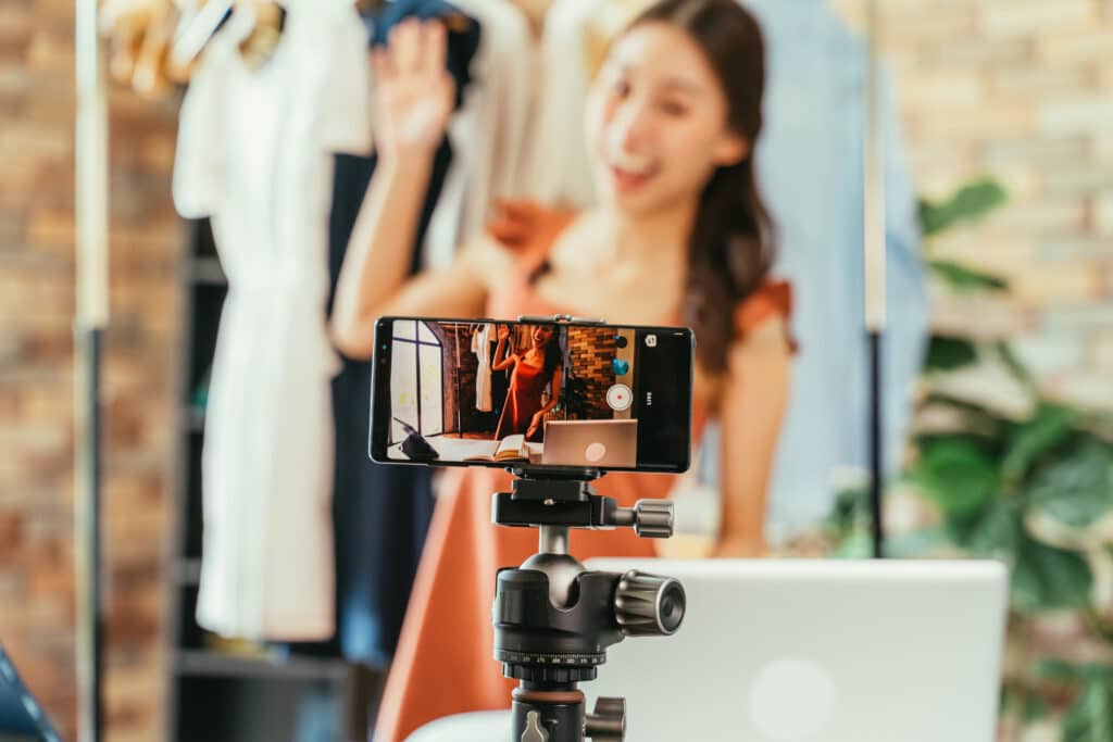 Young and beautiful Asian woman blogger showing clothes in front of smartphone camera while recording vlog video and live streaming at her shop