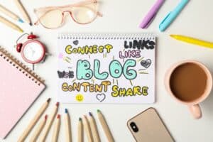A notebook with "Connect, Links, Like, Web, Blog, Content, Share" written in colorful letters sits among colored pencils, a mug, a phone, glasses, and an alarm clock—perfect for planning your blog strategy checklist.