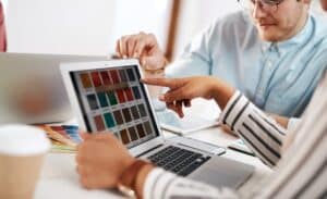 Two people review color swatches on a laptop screen at a desk, collaborating on a San Antonio Web Design project, with another laptop and color samples visible in the background.