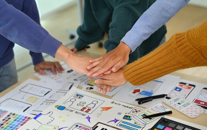Four people stack their hands over a desk covered with app design sketches, color swatches, and office supplies, reflecting the collaboration and creativity at the heart of San Antonio Web Design.