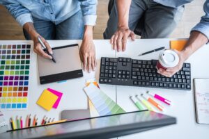 Two people work at a desk with a keyboard, drawing tablet, coffee cup, color swatches, pens, and notebooks—suggesting a collaborative San Antonio Web Design project.