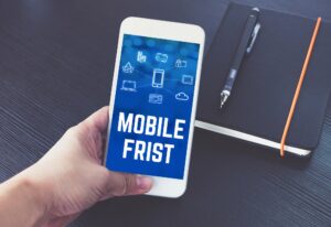 A hand holds a smartphone displaying the words "MOBILE FRIST" above icons, next to a closed black notebook with an orange pen on a dark surface—highlighting Website Design Mistakes San Antonio professionals should avoid.