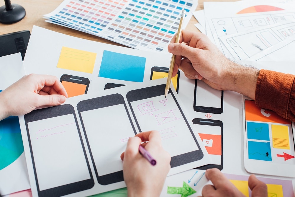 Designers sketching mobile phone wireframes on paper beside colour swatches