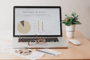 A laptop on a desk displays a monthly budget report with charts; nearby are a potted plant, eyeglasses, coins, papers on how to brief a web designer San Antonio, a pencil, and a wireless mouse.
