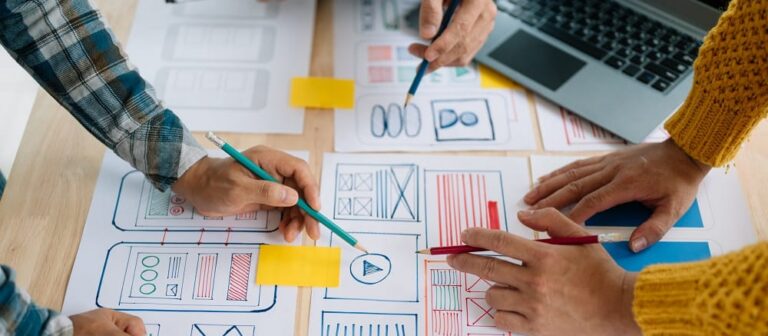 Three people's hands marking up wireframes and notes on a shared desk beside a laptop