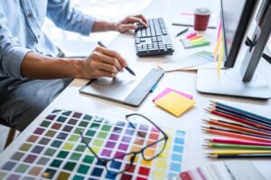 Person working at a desk with a graphics tablet, computer keyboard, color swatches, colored pencils, sticky notes, and a monitor—perfect for tackling projects like “How Long Does Web Design Take in San Antonio.”.