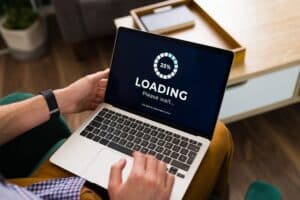 Person holding a laptop on their lap with a loading screen displaying "25% LOADING Please wait..." in white text and a progress circle graphic.