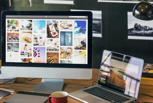 A desktop computer and a laptop on a wooden desk display photo galleries that showcase top website features; a mug, lamp, and photos on the wall are also visible.
