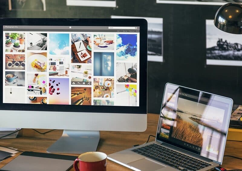 A desktop computer and a laptop on a wooden desk display photo galleries that showcase top website features; a mug, lamp, and photos on the wall are also visible.