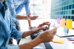 Two people discussing how to build a business website in San Antonio, with one typing on a keyboard and the other gesturing at the screen. Sticky notes are attached to the monitor.