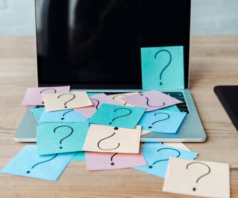 A laptop covered with colorful sticky notes, each displaying a large question mark, sits on a wooden desk next to notebooks—capturing the feeling of tackling website design questions.