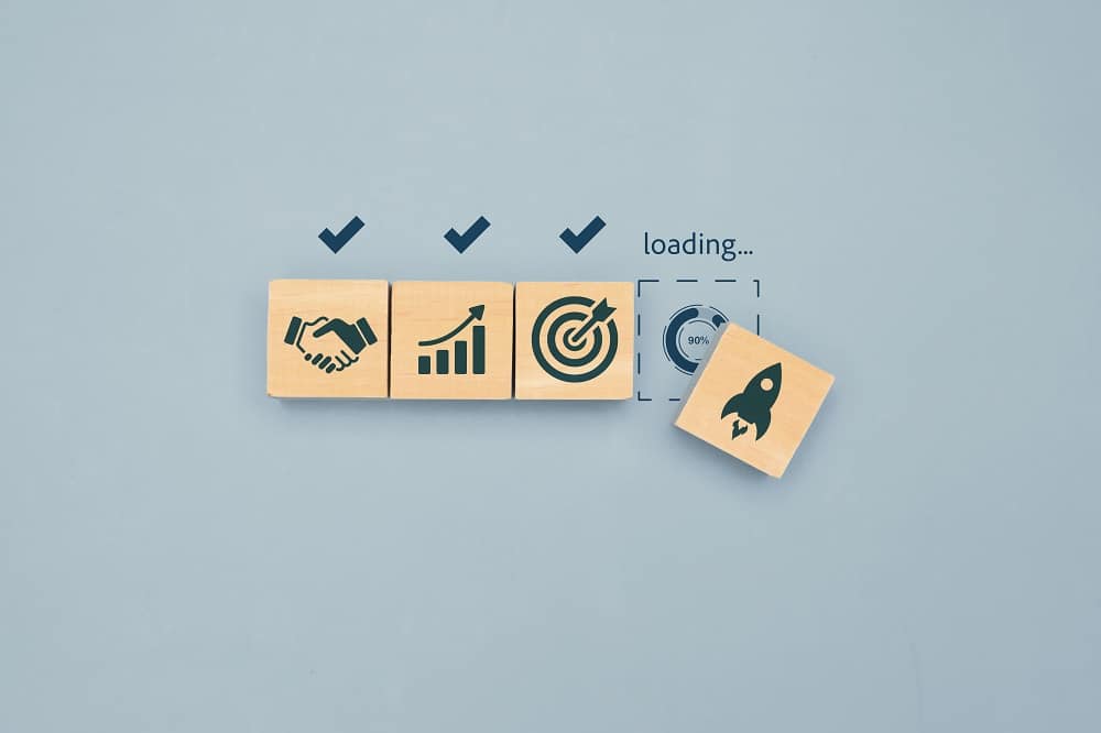 Four wooden cubes on a gray background—three with business icons and checkmarks, the fourth with a rocket icon and a "loading" progress bar at 90%—capture the anticipation of project launch, echoing the question: How Long Does Web Design Take in San Antonio?.