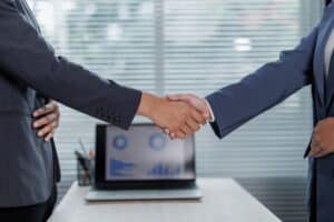 Two people in business attire shake hands in an office, with a laptop displaying charts on a desk—reflecting the collaborative spirit of a leading Web Agency in San Antonio.