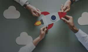 Three people hold a paper rocket cutout together on a table with paper clouds around it.