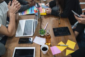 A group collaborates at a wooden table, using laptops, tablets, phones, color swatches, and sticky notes—with coffee and a beer bottle—immersed in the Web Design Process in San Antonio.