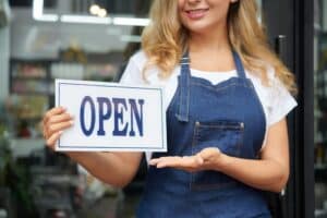 A woman wearing a denim apron holds an "OPEN" sign while standing at the entrance of a store, ready to welcome customers—discover how San Antonio businesses can use SEO to show up in Google AI Overviews and boost foot traffic.