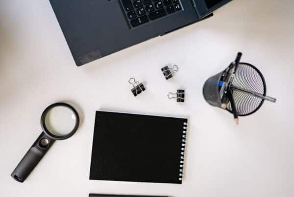 Overhead flat lay of a magnifying glass, blank black notepad, binder clips, and a laptop edge on a white desk.