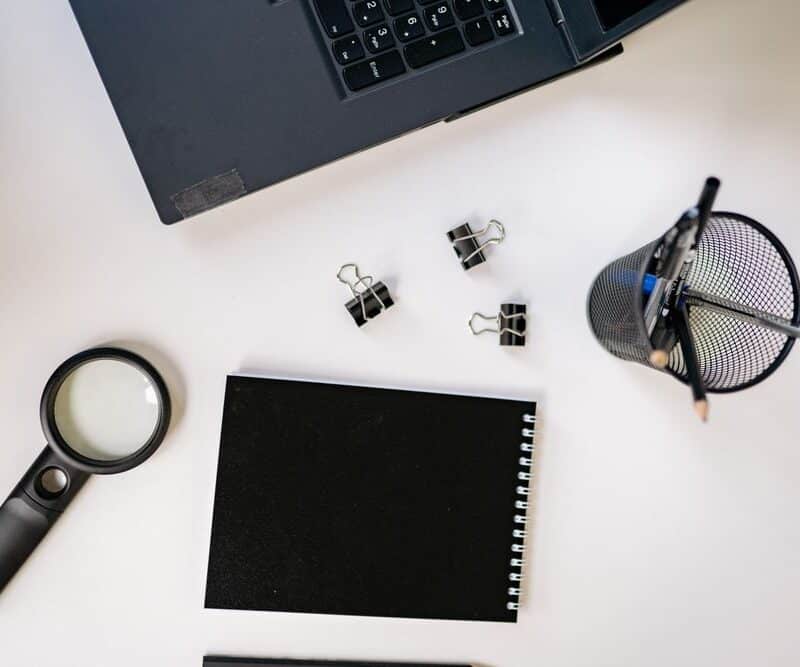 Overhead flat lay of a magnifying glass, blank black notepad, binder clips, and a laptop edge on a white desk.