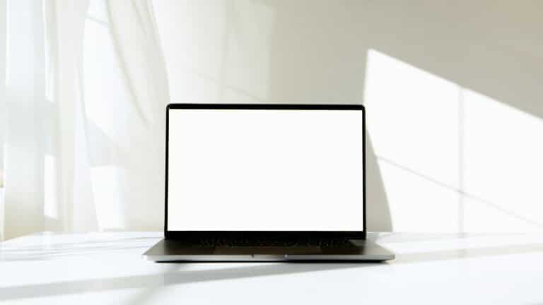 A laptop with a blank white screen on a bright white table beside a sunlit curtain, ready for a landing page design.