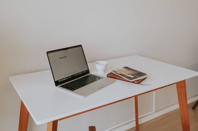 A minimal white desk holding an open laptop, a white coffee cup, and a closed notebook against a plain wall.