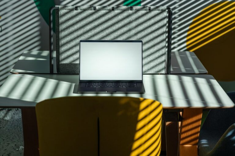 A laptop with a blank screen on a desk in a modern office, lit by striped daylight from window blinds.