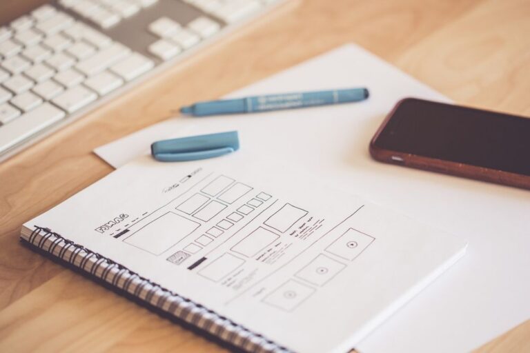 A spiral notebook showing a hand-drawn website layout wireframe beside a keyboard and smartphone on a light wooden desk.
