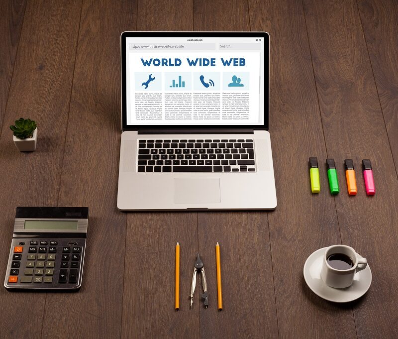 A laptop displaying “World Wide Web,” a calculator, compass, pencils, markers, a cup of coffee, and a small potted plant arranged neatly on a wooden desk.