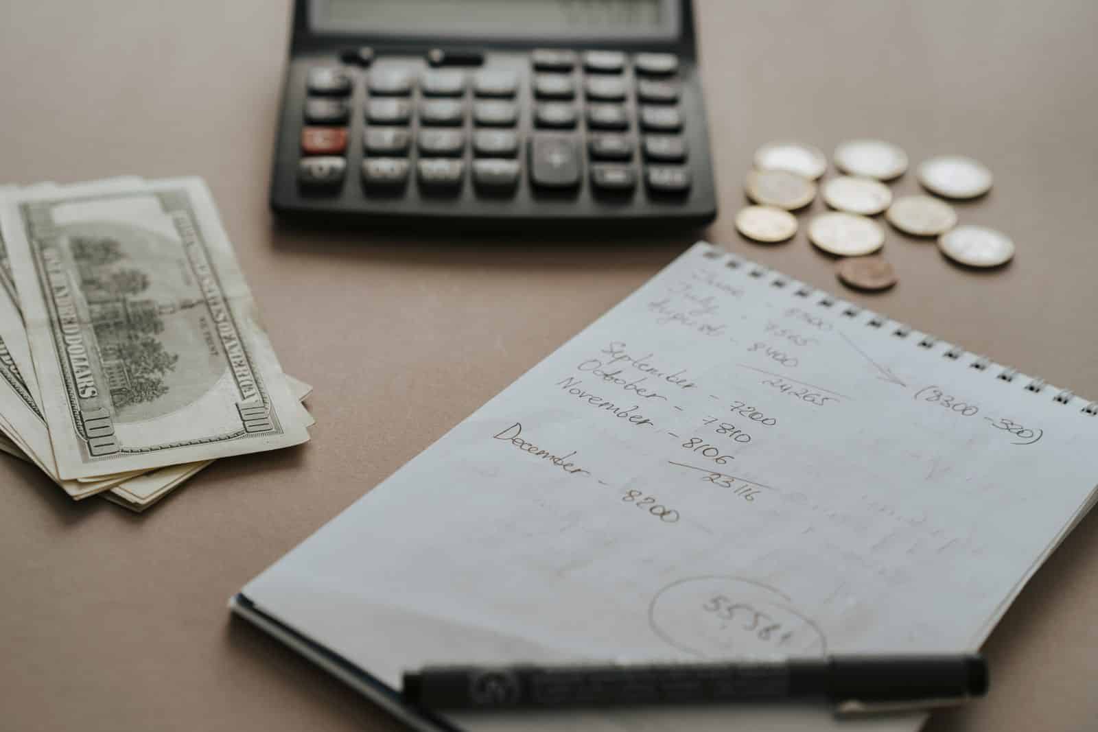 Calculator, dollar bills, coins, and a notebook of monthly figures on a desk