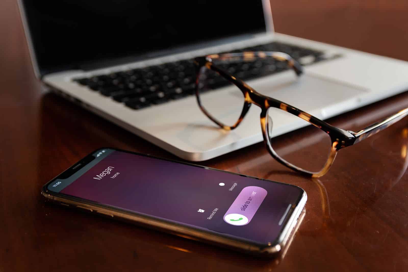 Smartphone on a desk showing an incoming call next to a laptop and eyeglasses