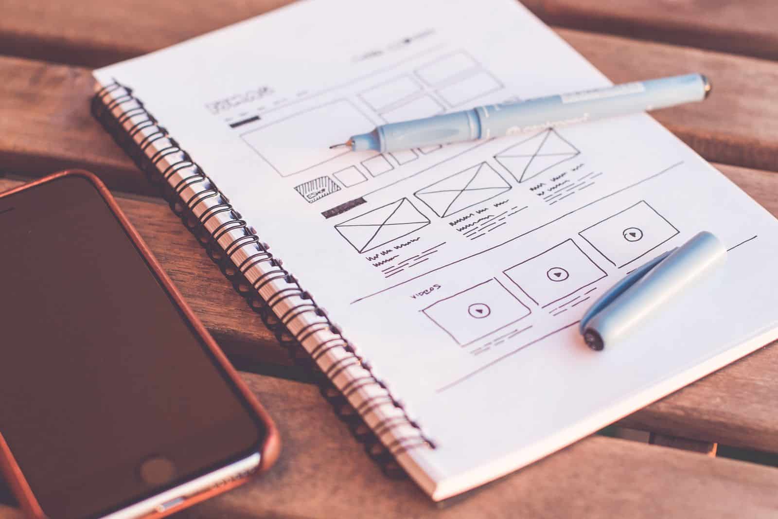 Notebook with hand-drawn website wireframe sketches next to a smartphone and pen